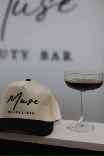 Muse Beauty Bar embroidered cap beside a coupe glass inside the salon